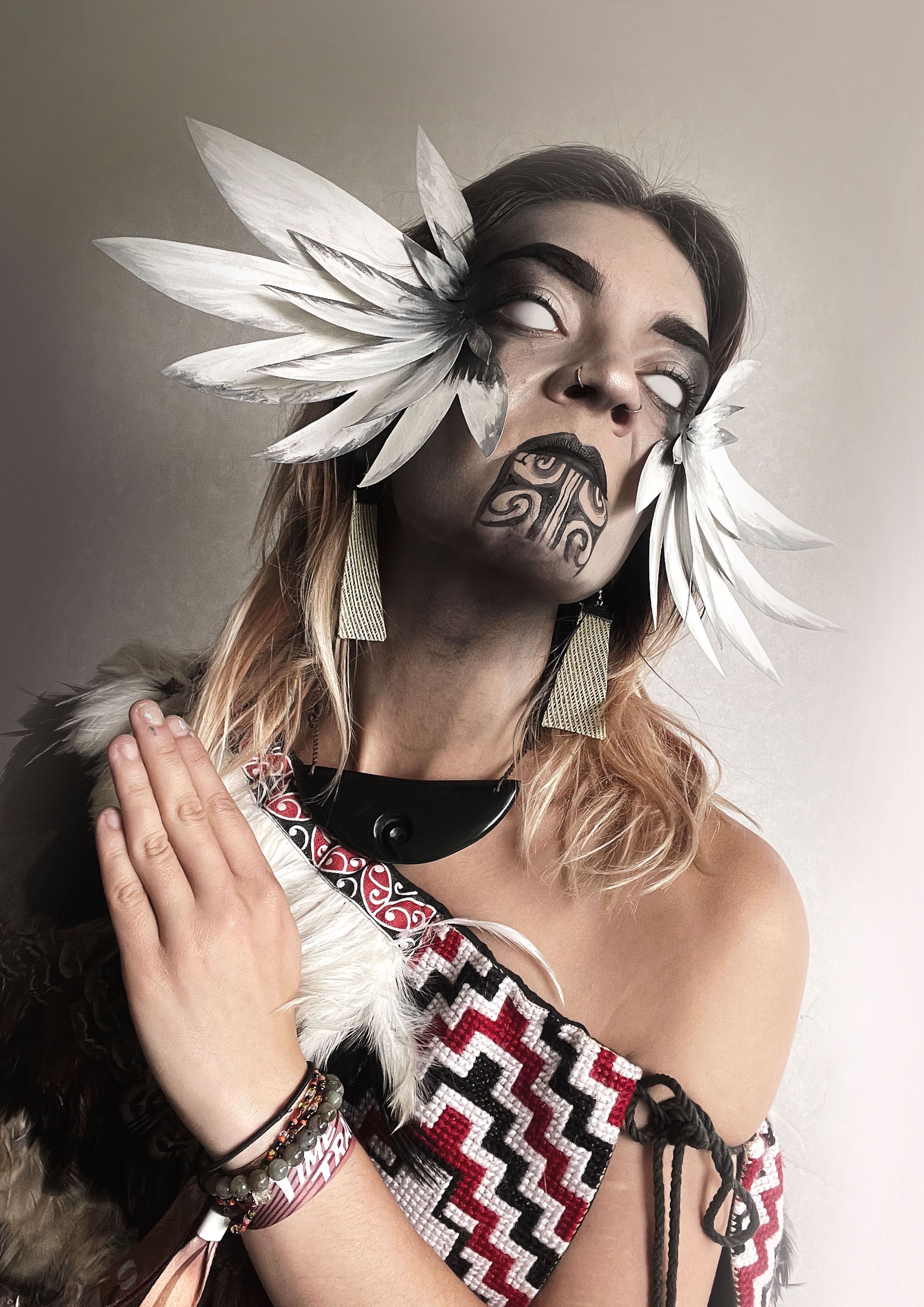A femme person in a woven kahu/cloak, hand raised in a wiri in front of them, eyes painted over with white and with a white headpiece of feather-like shapes